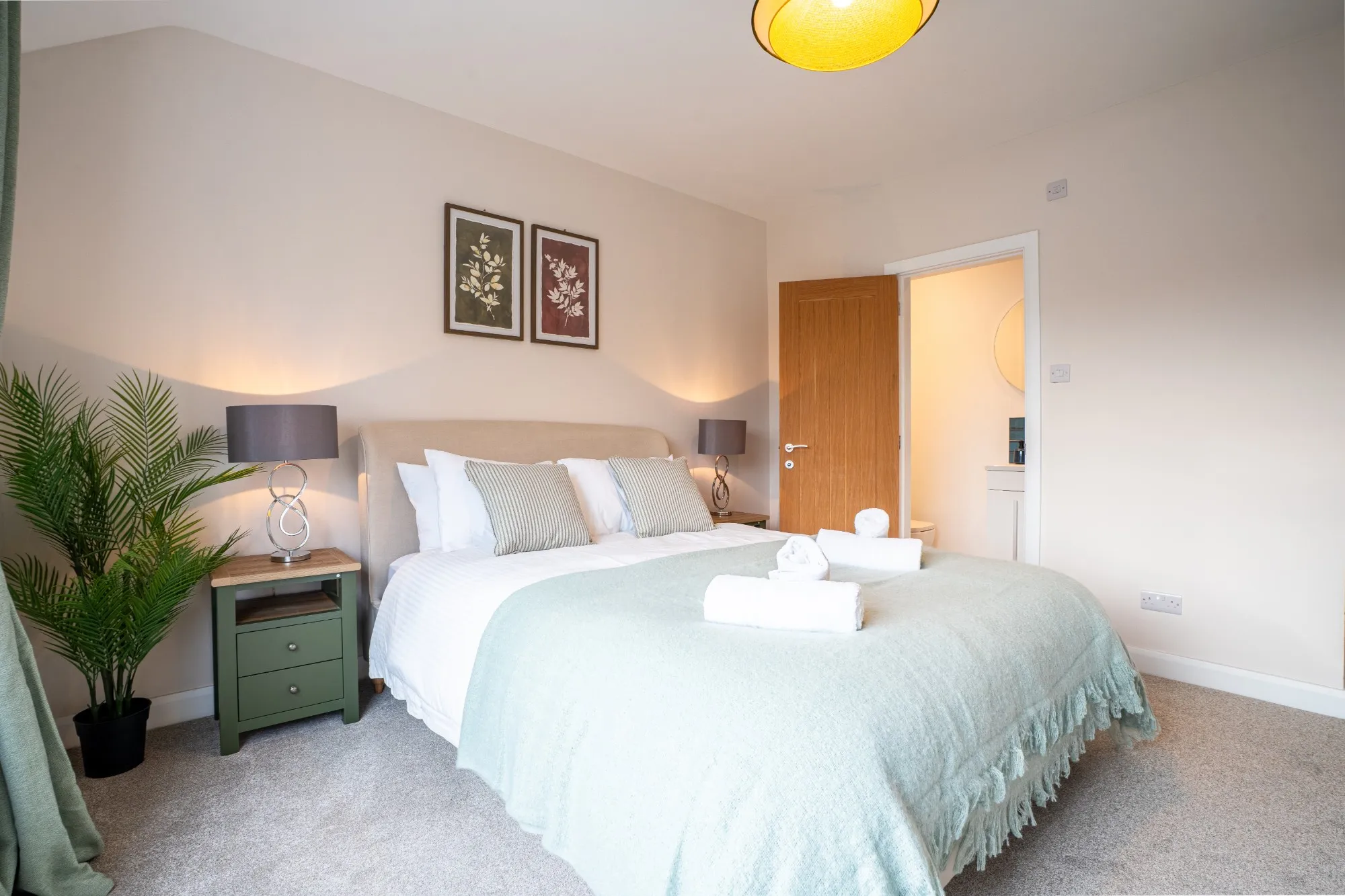 A double bedroom with an upholstered headboard, sage throw, green bedside tables and an en suite through the oak door